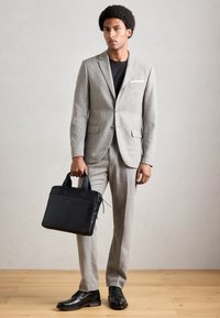 Mann iført lys grå dress og sorte sko, som holder en svart koffert, står på tregulv med nøytral bakgrunn.