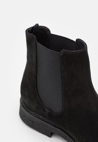 Botte Chelsea en daim noir avec panneau latéral élastique et languette de tirage, dotée d'une semelle texturée et d'un design à bout arrondi.