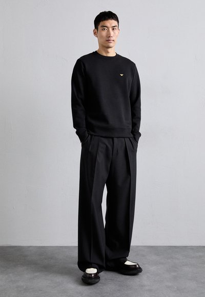 Black sweatshirt with a ribbed hem and cuffs, featuring a small gold logo. Paired with wide-leg black trousers and black footwear with white accents.