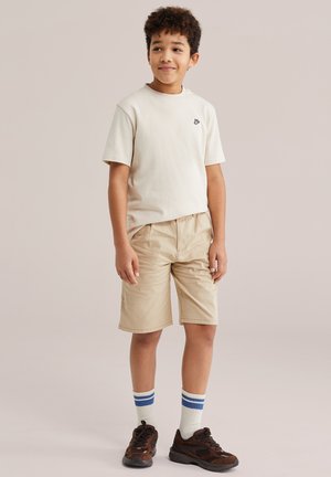 Niño con cabello rizado, vistiendo pantalones cortos beige, una camiseta crema con un pequeño emblema, calcetines blancos con rayas azules y zapatillas oscuras, de pie relajado.