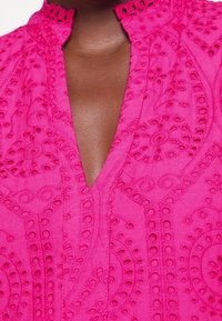 Fuchsia blus med intrikata broderier, som har en V-ringning och dekorativa utskärningar längs kragen och frontpanelen. Slät textur.