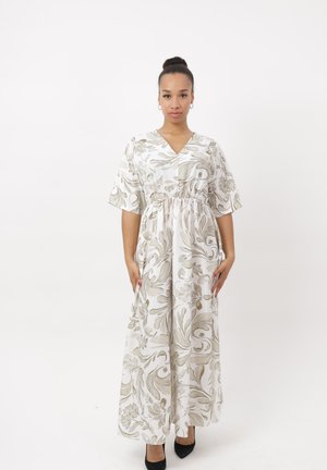 Vrouw staand, draag een witte maxi-jurk met beige bloemmotieven, korte mouwen en zwarte puntige schoenen op een effen witte achtergrond.