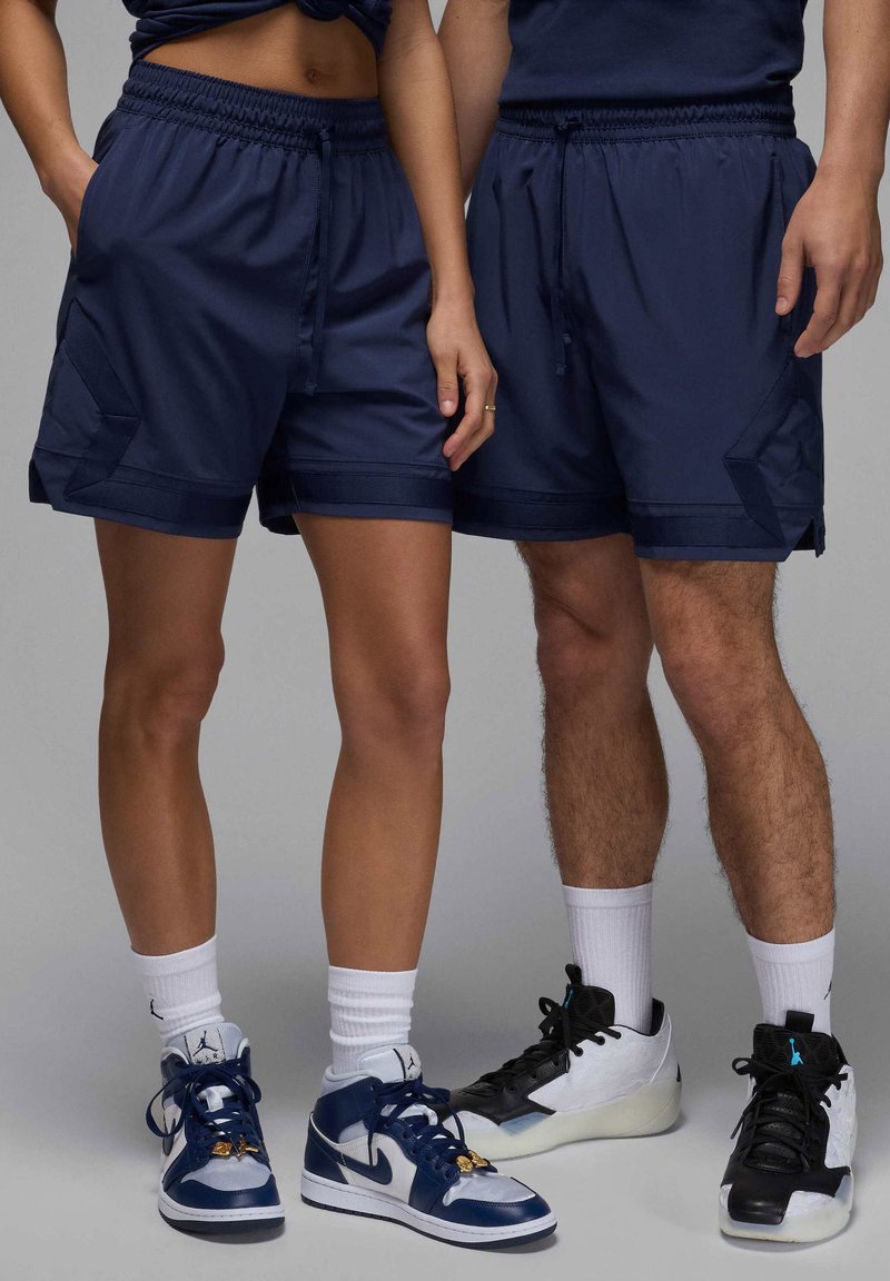 Pantaloni sportivi navy con cintura elastica, tasche laterali e dettagli a contrasto. Indossati con sneakers alte e calze bianche.