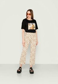 T-shirt gráfica preta com a impressão "LAZY DAY", combinada com umas calças bege às riscas de zebra. O modelo usa óculos de sol laranja e sandálias pretas grossas.