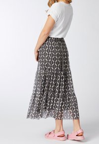 Mujer vestida con una blusa blanca de manga corta, una larga falda estampada y sandalias rosas, de pie frente a un fondo liso.