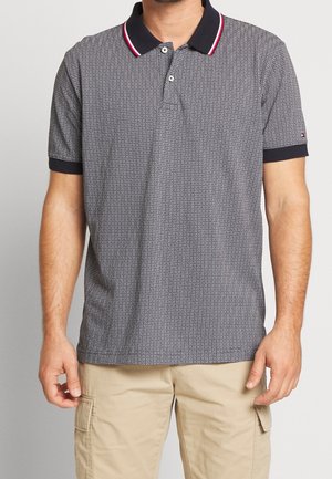 Mann trägt ein kurzärmeliges, marineblau-weiß gemustertes Poloshirt mit rot gestreiftem Kragen und beigen Cargo-Shorts, steht vor einem einfarbigen Hintergrund.