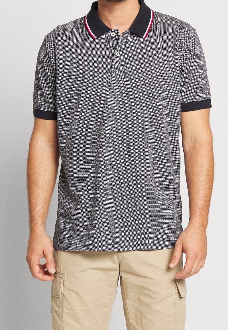 Homme portant un polo à manches courtes bleu marine et blanc à motifs, col rayé rouge, et un short cargo beige, debout devant un fond uni.