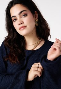 Marineblauwe gebreide trui met brede mouwen, gelaagde gouden en slangpatroon kettingen, grote gouden en zwarte oorringen en meerdere ringen.