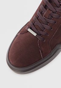 Zapatilla de ante burdeos con un acabado texturizado, cordones planos y suela de goma. Presenta un acento de logo metálico en la lengüeta.