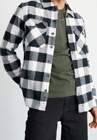 Camicia di flanella a quadretti bianco e nero con due tasche sul petto, chiusura a bottoni e colletto, indossata sopra una maglietta verde scuro e pantaloni neri.