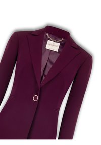 Bordeaux blazer met een gekartelde revers, een enkele knoop met gouden accent en subtiele schouderstructuur. Satijnen voering zichtbaar bij de kraag.