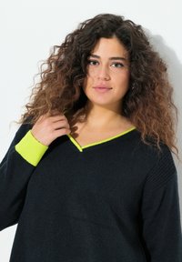 Svart tröja med V-ringning, med lysande limegrön kant vid halsringningen och ärmsluten. Mjuk textur och avslappnad passform är tydliga.