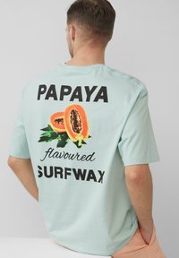 Jasnoniebieska bawełniana koszulka z grafiką dwóch papai oraz czarnym napisem "WOSK DO SURFERÓW o smaku PAPAI" na plecach.