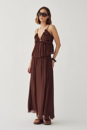 Žena v hnedých maxi šatách bez rukávov s volánmi, nadrozmerných čiernych slnečných okuliaroch, čierno-bielych náramkoch a platformových sandáloch stojaca pred jednoduchým pozadím.