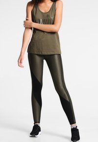 Top sin mangas verde oliva combinado con leggings negros y estampados. Los leggings presentan una textura similar a malla y acentos negros en contraste. Zapatillas negras.