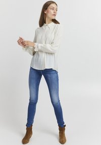 Helle, cremefarbene Bluse mit Spitzenkragen und Spitzenakzenten an den Ärmeln, kombiniert mit blauen Skinny-Jeans und braunen Stiefeletten.