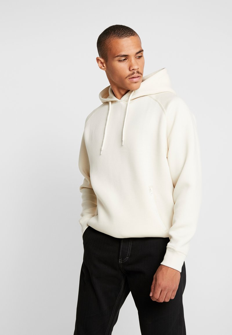 Hoodie color beige chiaro con cappuccio regolabile, tasca a marsupio e maniche raglan. Jeans neri con dettagli di cucitura visibili. Design semplice e minimalista.