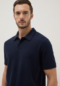 Hombre con cabello corto y barba, vistiendo una camisa polo azul marino de manga corta, mirando hacia adelante sobre un fondo claro y sencillo.