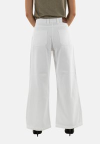 Pantalones de pierna ancha blancos con una cintura alta, que cuentan con dos bolsillos traseros y una etiqueta de cuero marrón. Textura suave y diseño limpio.