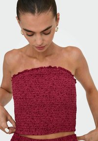 Bordeaux strapless top med tekstureret, flæset kant, tætsiddende design og strækbart stof, båret af en model mod en lys baggrund.