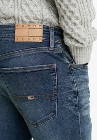 Jeans dżinsowe w ciemnoniebieskim kolorze z teksturowanym wykończeniem. Posiadają skórzaną metkę na pasa z wytłoczonym napisem "TOMMY JEANS". Maleńki, kolorowy detal w postaci flagi na kieszeni.