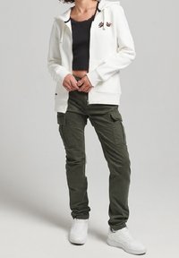 Sudadera blanca con cremallera y bolsillo frontal, top negro corto, pantalones cargo verde oliva con bolsillos laterales y zapatillas blancas.