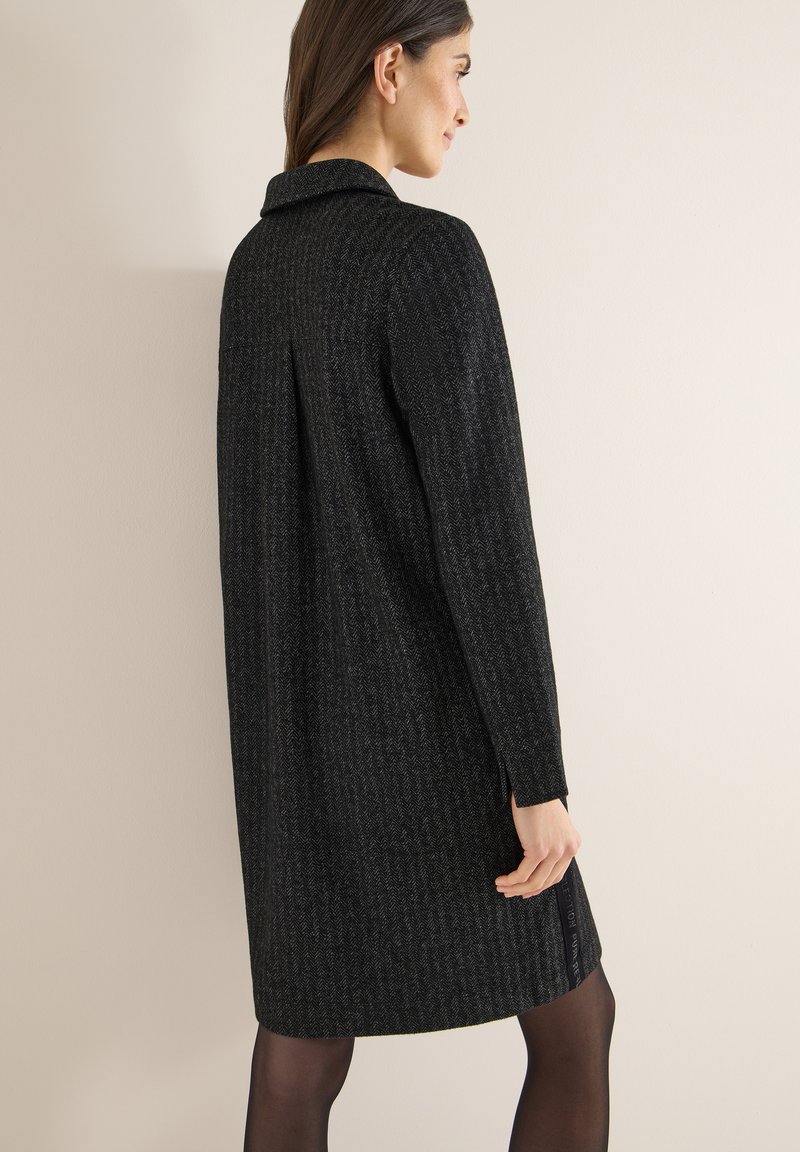 Vestido negro con patrón de espiga, mangas largas, cuello y aberturas laterales, hecho de tela texturizada, que llega hasta la mitad del muslo.