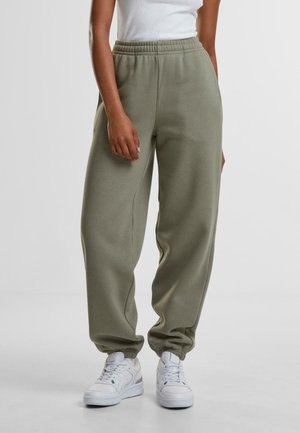 Stående person iført olivengrønne joggingbukser med elastiske manchetter, hvide sneakers og en hvid ærmeløs top mod en ensfarvet baggrund.