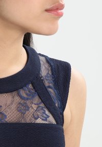 Mörkblå klänning med en texturerad tyg och blommig spetspanel vid halsringningen och axlarna, som visar upp intrikata mönster.