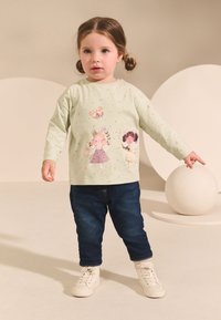 Svetlozelená košeľa s dlhým rukávom s potlačou detských ilustrácií, v kombinácii s tmavomodrými džínsami a bielymi teniskami. Minimalistické pozadie.