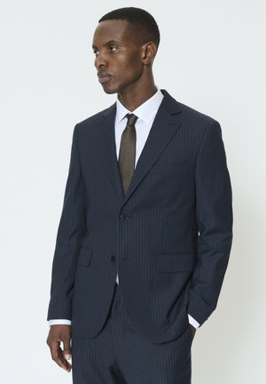 Marineblå dragt med nålestriber, to knapper, notch-lapel revers og bukser med flad front. Hvid skjorte og brunt tekstureret slips fuldender looket.