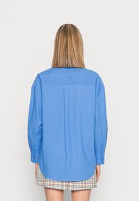 Camicia azzurra chiara a maniche lunghe con piega sul retro e polsini abbottonati, realizzata in un tessuto liscio, abbinata a una gonna a quadri.