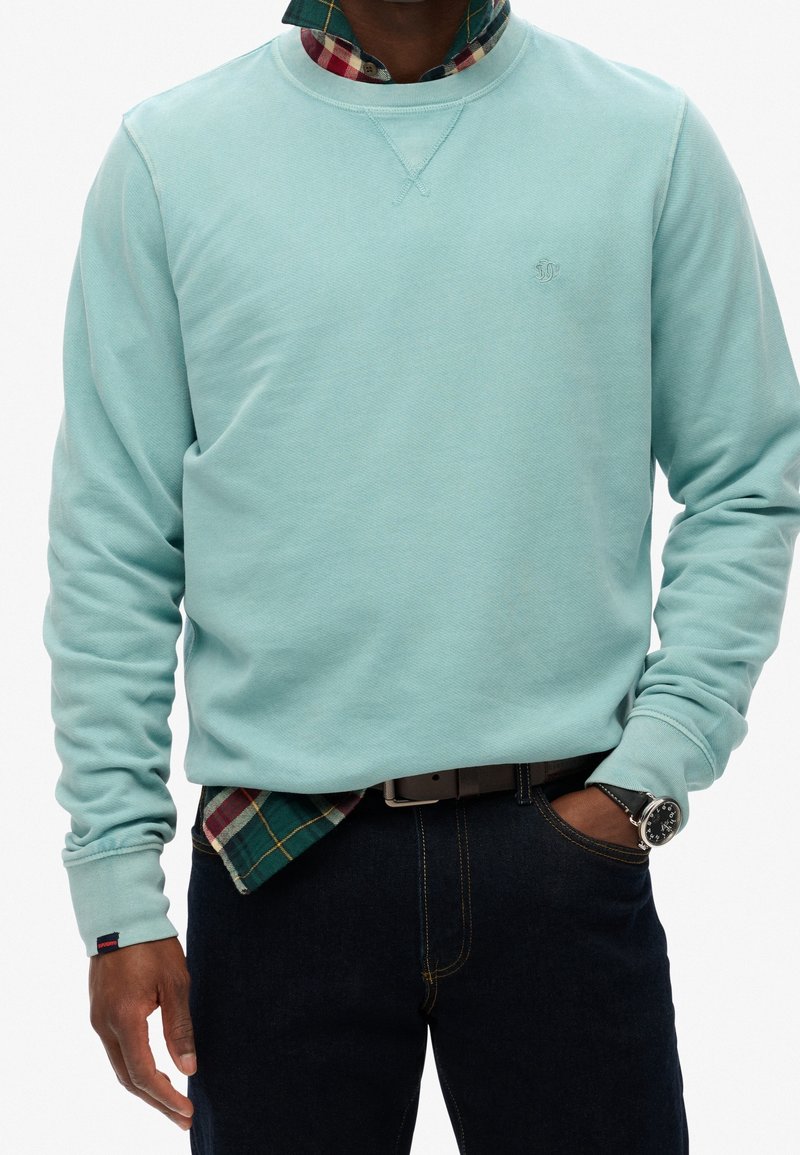 Homme portant un sweat-shirt bleu clair par-dessus une chemise à carreaux verts et rouges, un jean foncé, une ceinture noire et une montre noire avec un boîtier argenté.