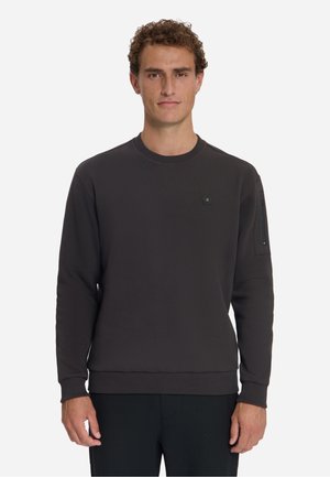 Man draagt een effen donker sweatshirt met lange mouwen en ronde hals, met een klein logo op de borst en een ritsvak aan de linker mouw.