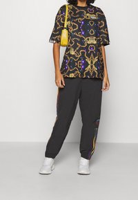 Svart mönstrad t-shirt med guldhalsband och färgglada accenter, kombinerad med svarta joggers med sidostreck i flera färger och vita sneakers.