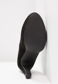 Chaussure à talon haut en suède noir avec un bout pointu, une texture lisse et une silhouette élégante et courbée. La semelle est en caoutchouc noir texturé.