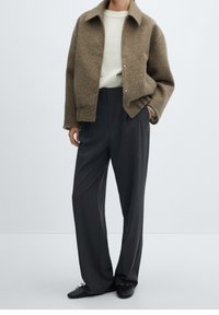 Brown fuzzy jacket with a collared neck, cream sweater underneath, and grey wide-leg trousers. Black shoes with laces are worn.