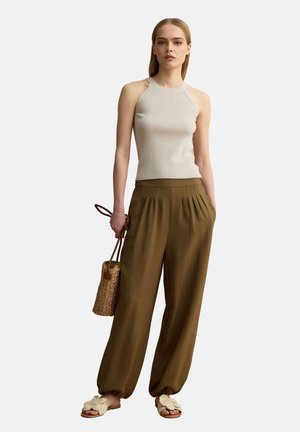 Frau trägt beige Neckholder-Top, lockere braune Hose, hält geflochtene Handtasche und trägt Slip-On-Sandalen mit Schleifendetail, steht vor weißem Hintergrund.