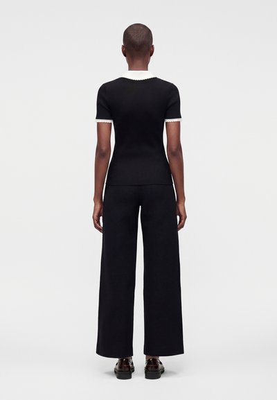 Personne vue de dos portant un haut noir à manches courtes avec un col blanc et un pantalon noir à jambes larges sur un fond clair uni.