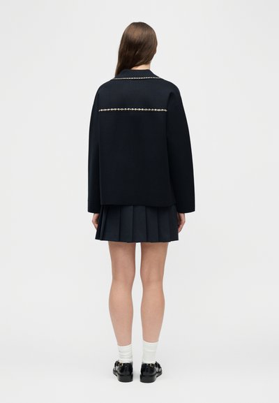 Γυναίκα που στέκεται με την πλάτη, φορώντας μαύρο μπουφάν με διακόσμηση από πέρλες, πλισέ μαύρη φούστα, λευκές κάλτσες και μαύρα μοκασίνια με χρυσές λεπτομέρειες.