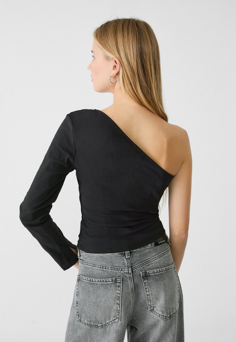 Top nero con spalla scoprata, dal design aderente e manica lunga, realizzato in tessuto morbido. Abbinato a jeans grigi a vita alta.