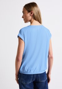 Lichtblauwe blouse met korte mouwen, ronde halslijn en elastische zoom. Glad stof, subtiele glans en opgerolde mouwen. Draagt met donkerblauwe jeans.