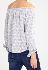 Blusa senza spalle di colore bianco con motivo fogliare blu navy, caratterizzata da dettagli arricciati sul collo e maniche legate. Abbinata a jeans scuri.