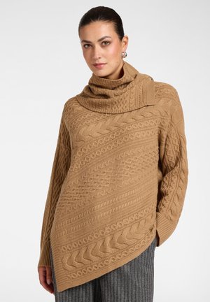 Vrouw draagt een lichtbruine kabelgebreide asymmetrische poncho trui met een hoge opgevouwen kraag en grijze geribbelde broek.
