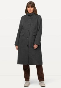 Lungo cappotto di lana grigio con colletto alto, chiusura a bottoni e tasche laterali. È abbinato a pantaloni marroni e sneakers di colore chiaro.