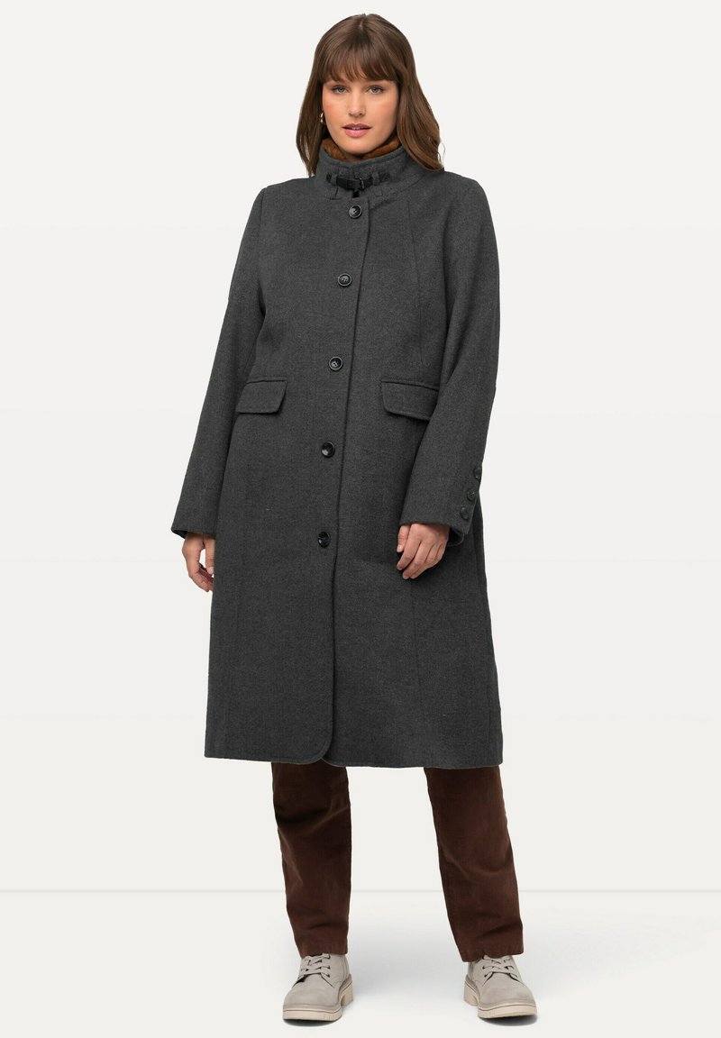 Lungo cappotto di lana grigio con colletto alto, chiusura a bottoni e tasche laterali. È abbinato a pantaloni marroni e sneakers di colore chiaro.