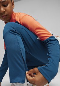 Pantalones deportivos azules con un acabado texturizado y una camiseta de manga larga naranja. Los pantalones tienen un detalle de cremallera lateral. La persona está ajustando el dobladillo.