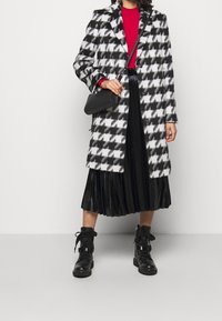 Houndstooth-mønstret frakke i sort og hvid, parret med en rød ribbet sweater, sort plisseret nederdel, sorte ankelstøvler og en lille sort taske.