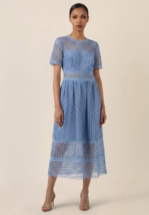 Frau steht vor einem einfarbigen beigefarbenen Hintergrund, sie trägt ein hellblaues Spitzen-Midikleid mit kurzen Ärmeln und hohe Absätze.