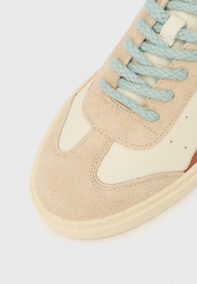 Beige suède en crèmekleurige leren sneaker met lichtblauwe veters, ronde neus en contrasterende rode accent aan de zijkant. Gestructureerd oppervlakpatroon.